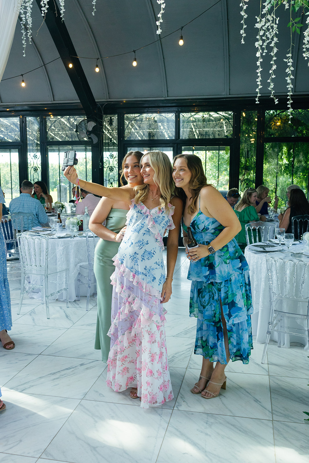 Three women in floral dresses pose for a selfie, smiling as they enjoy the celebration.