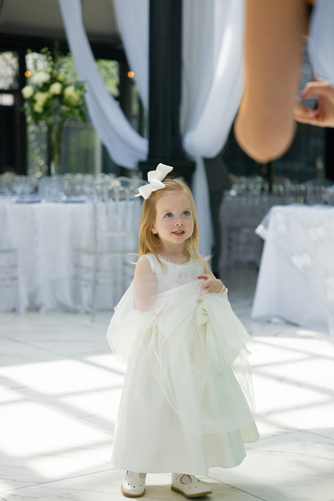 A young flower girl in a white dress and big bow looks up sweetly while standing inside the decorated reception space.