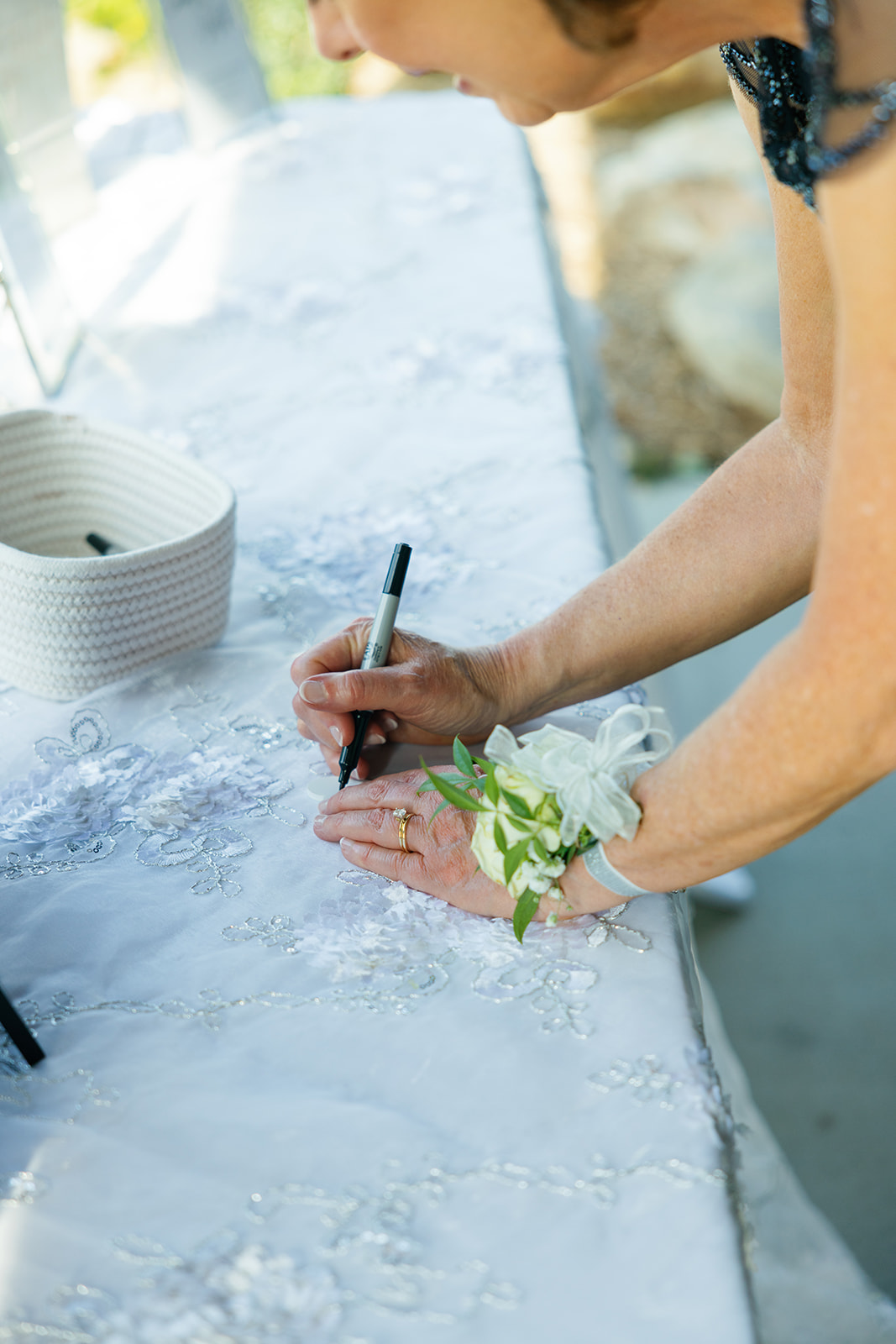 A close-up of a woman’s hand with a corsage writing on a white embroidered cloth guestbook with a black marker.