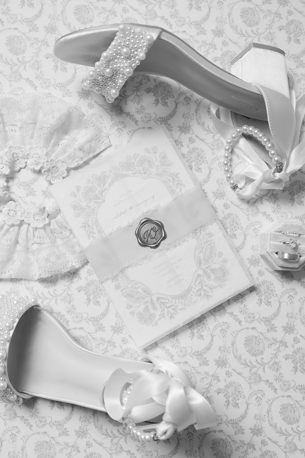 black and white photo featuring pearl-embellished bridal heels, a lace garter, pearl bracelet, wedding rings in a box, and an invitation with a wax seal.