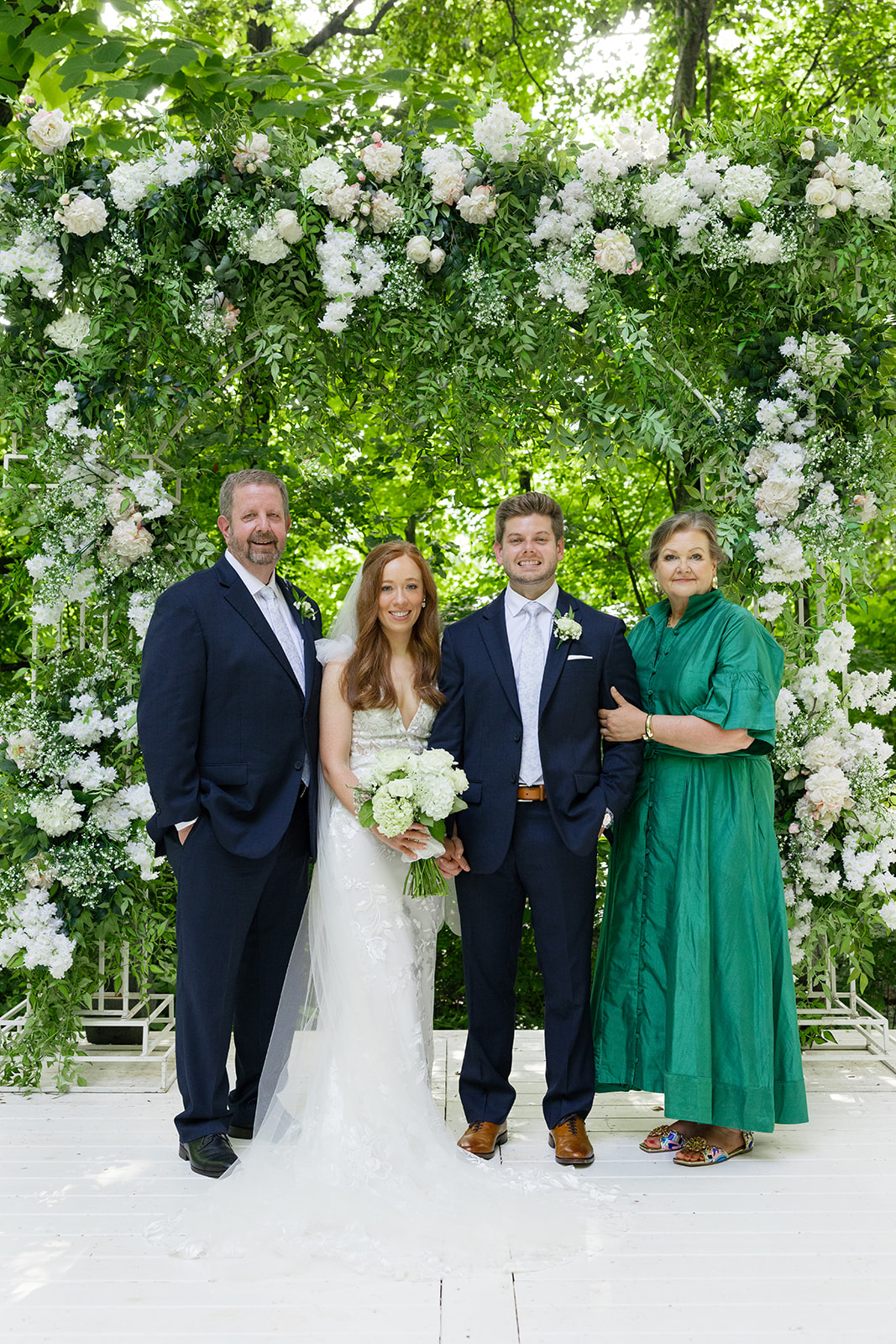 Bride and groom pose with groom’s parents under a floral ceremony arch.