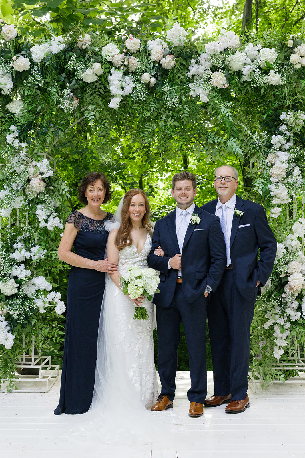Bride and groom pose with bride’s parents under the ceremony arch.