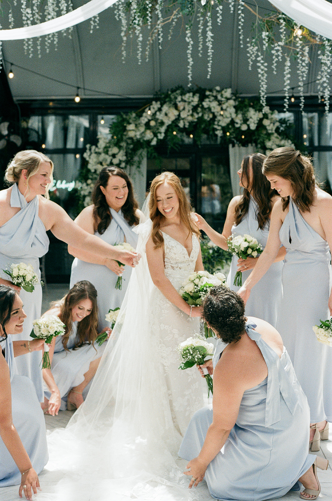Bride smiles as her bridesmaids in light blue dresses gather around, adjusting her veil and holding white bouquets inside the reception space.