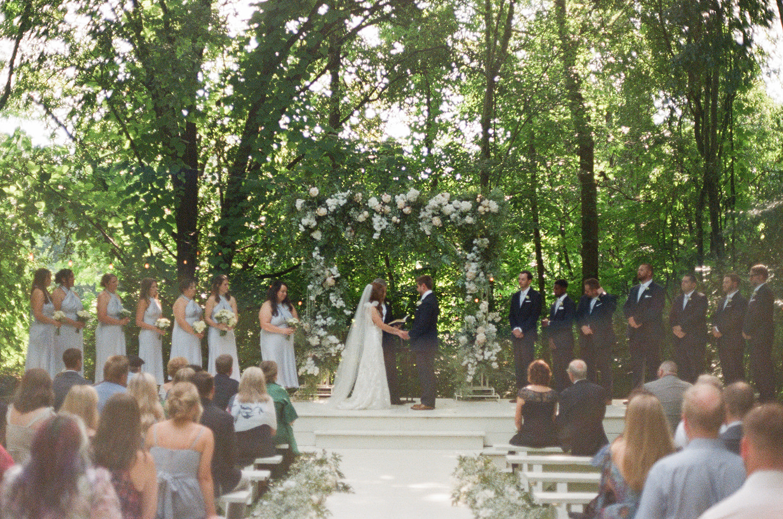 Bride and groom stand under a lush floral arch, exchanging vows during their outdoor ceremony, surrounded by bridesmaids and groomsmen as guests watch.