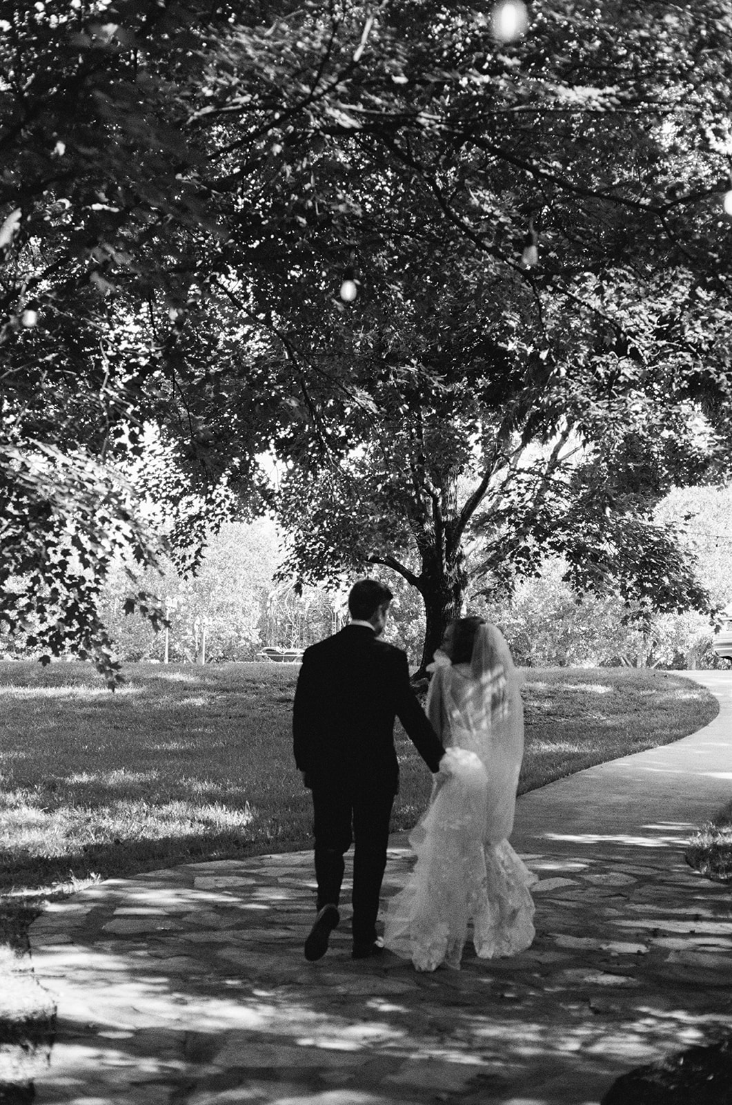 Black-and-white photo of bride and groom walking hand in hand down a stone path beneath tall trees after their ceremony.