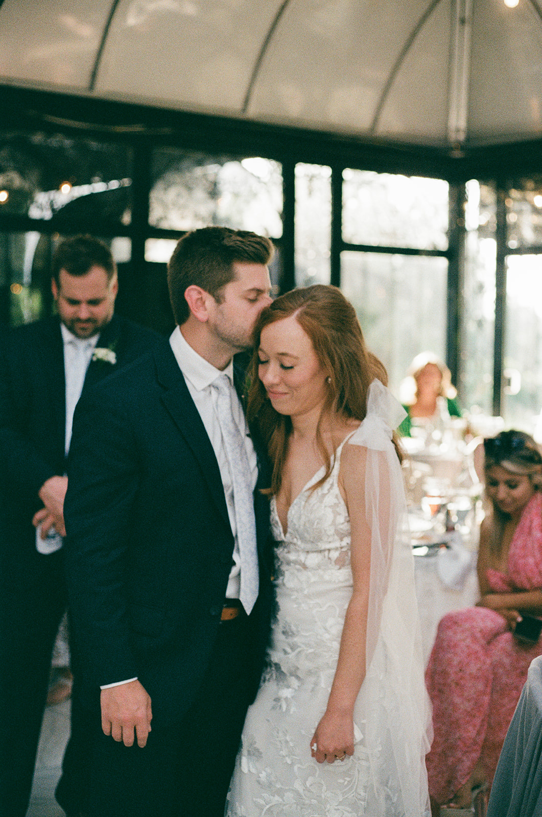 Groom in a navy suit gently kisses his bride’s forehead during their Greenville, SC wedding reception at Edinburgh West, a sweet, candid moment captured by a Greenville wedding photographer.