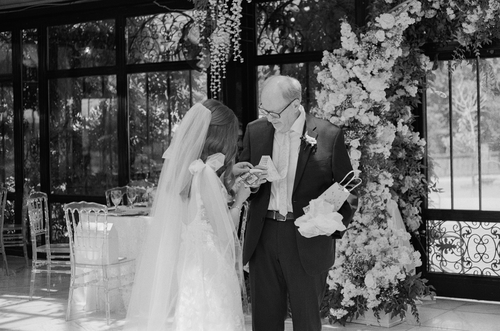 Bride in lace gown shares a joyful moment with her father as they exchange a gift inside the floral-decorated reception space.