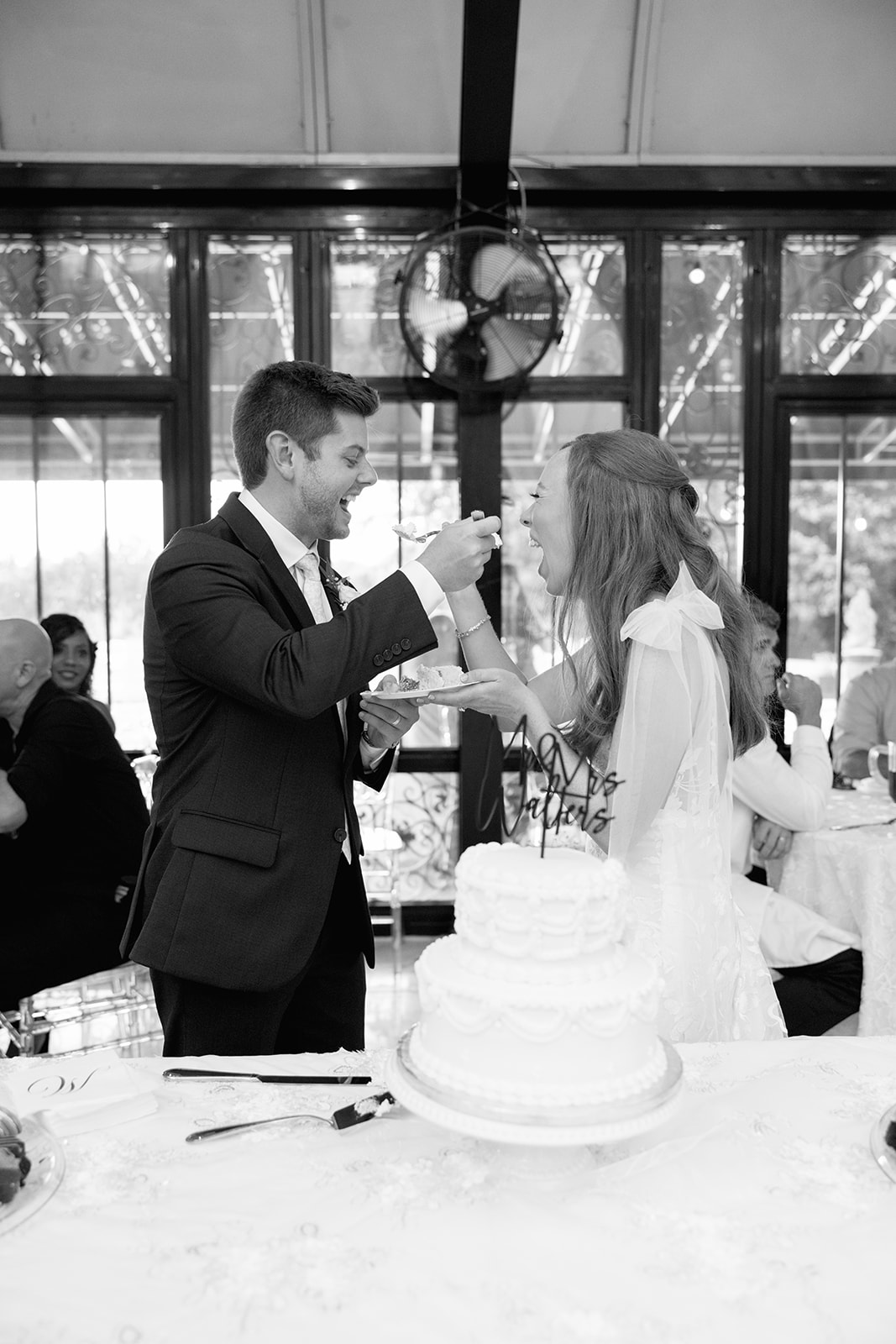 Black-and-white photo of the groom playfully feeding cake to the bride during the cake-cutting tradition.