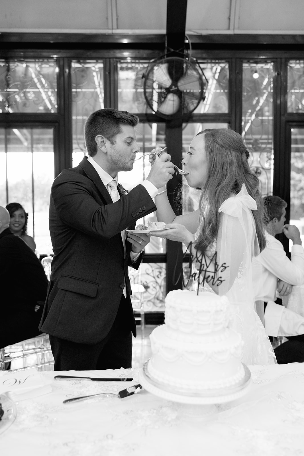 Black-and-white photo of the bride and groom feeding each other cake at the same time, laughing together.