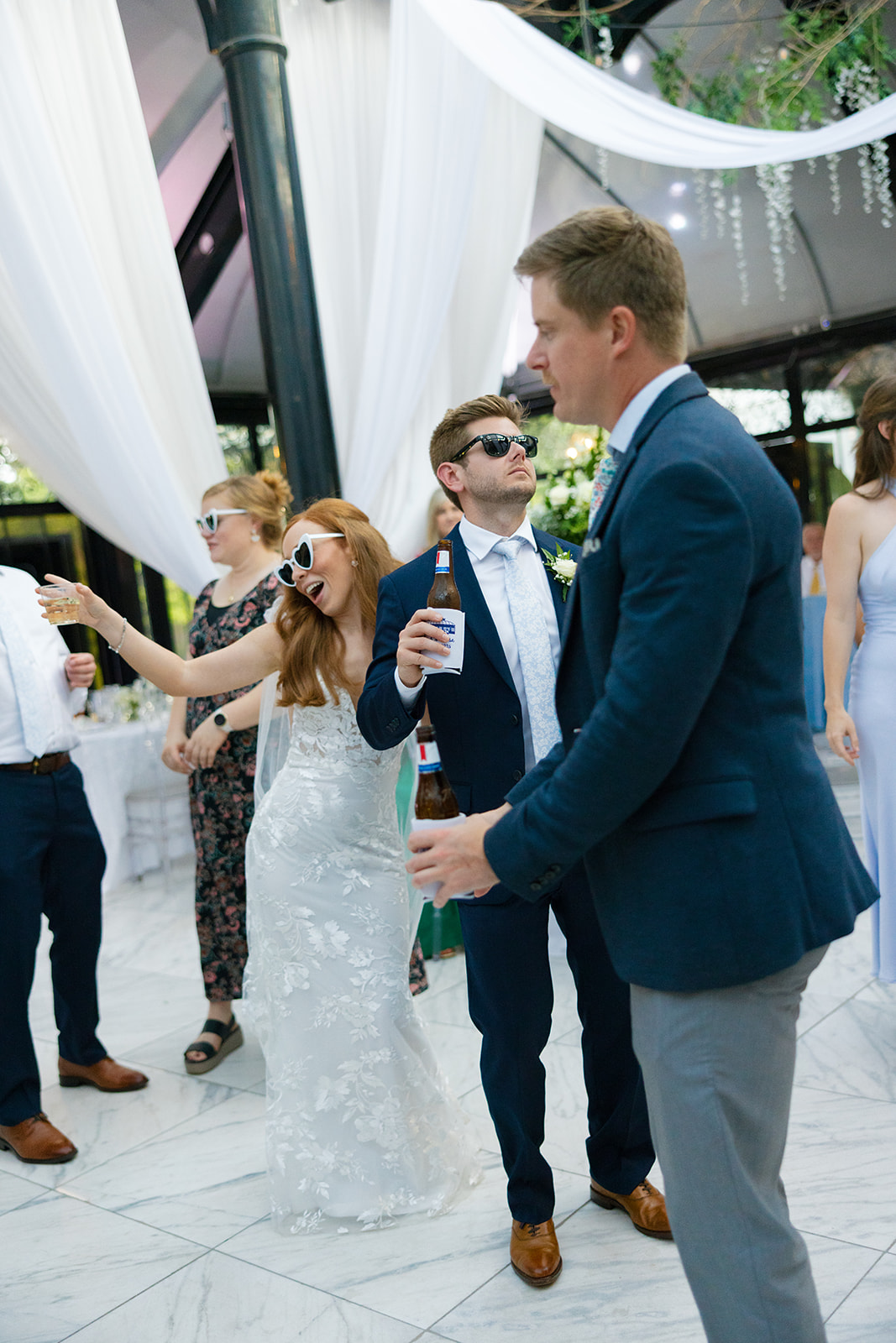 The bride and groom, both wearing heart-shaped sunglasses, dance and hold drinks as guests celebrate around them.
