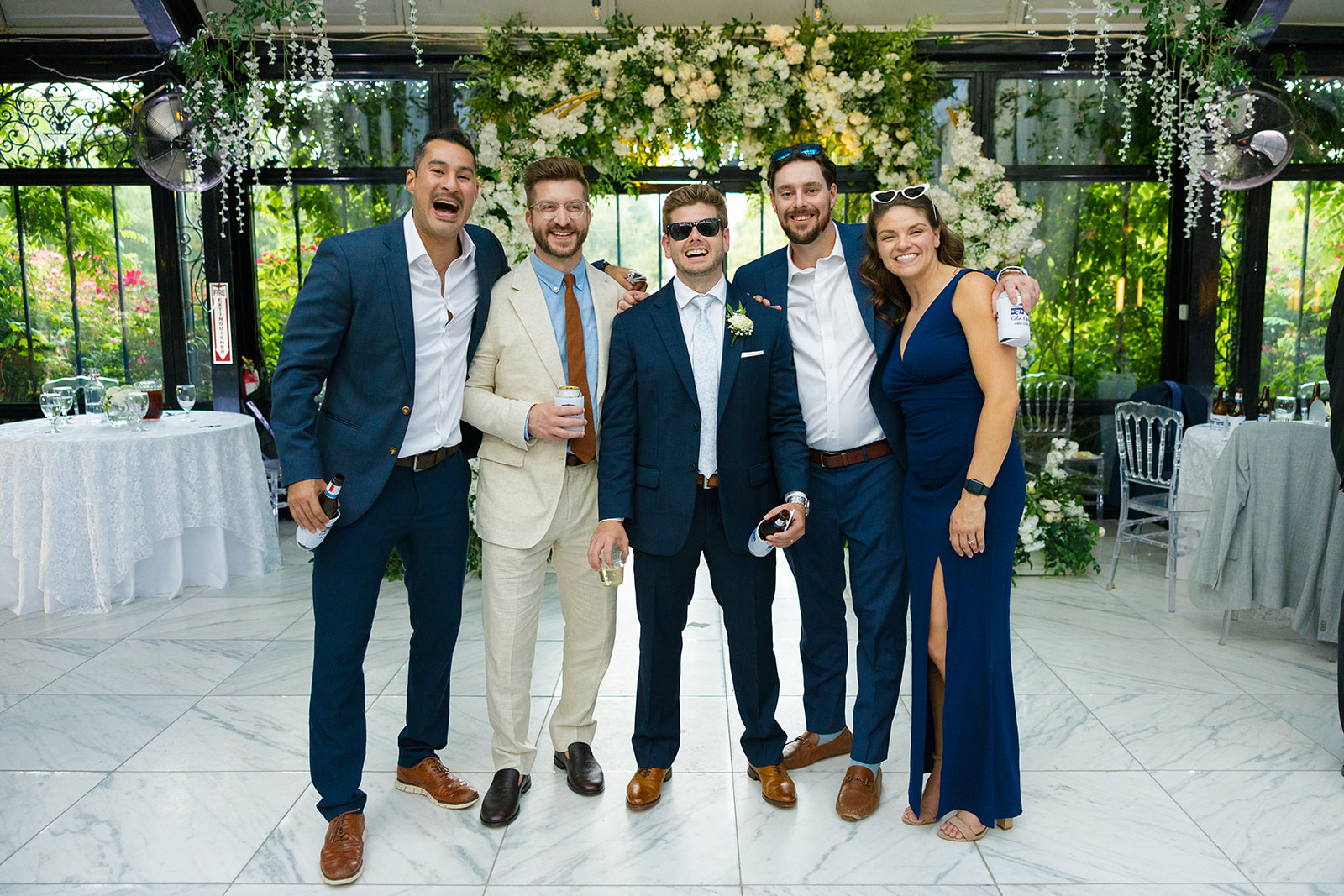 Groom in navy suit and sunglasses poses with four friends in suits and a blue dress, smiling together inside the reception venue decorated with white florals.