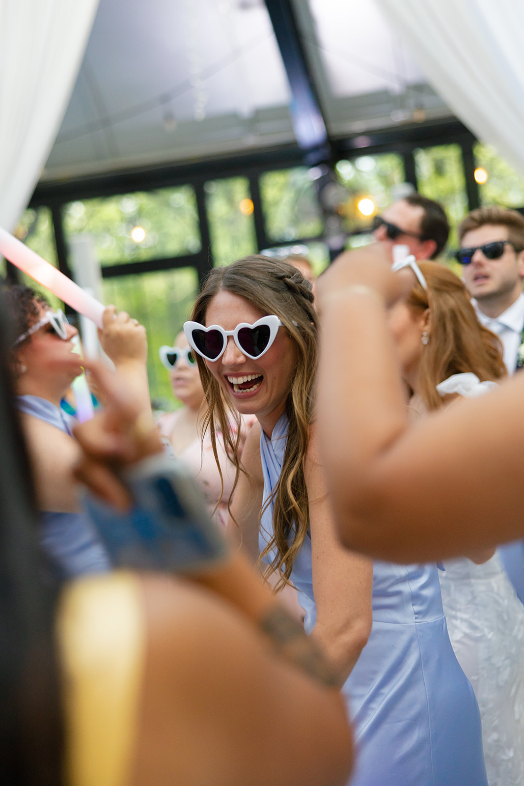 Bridesmaid in light blue dress dances with heart-shaped sunglasses on, laughing and surrounded by other guests on the reception dance floor.