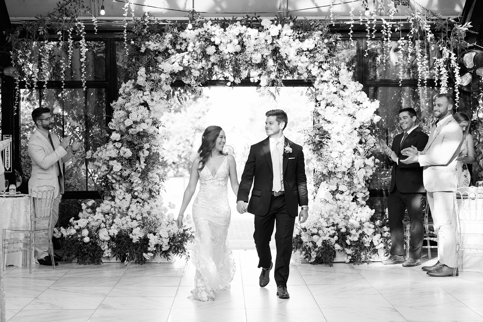 Bride and groom walk hand-in-hand into their reception under a floral arch as guests applaud.