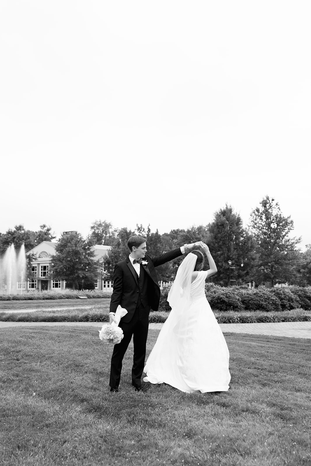 Groom twirling bride on a grassy lawn with a fountain and trees in the background – wedding planning advice: schedule relaxed outdoor portraits for natural, joyful moments.