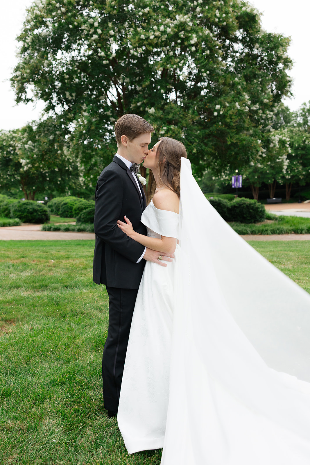 Groom kissing bride on a grassy lawn with trees in the background – wedding planning advice: schedule relaxed outdoor portraits for natural, joyful moments.