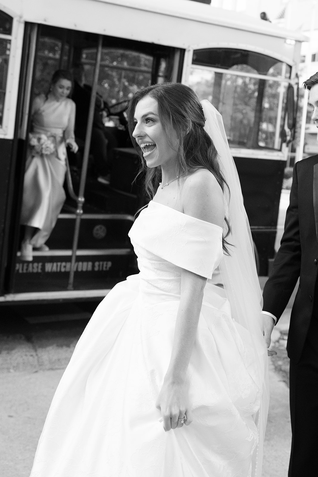 Black and white photo of bride beaming with excitement as she exits the trolley during her wedding day celebration.