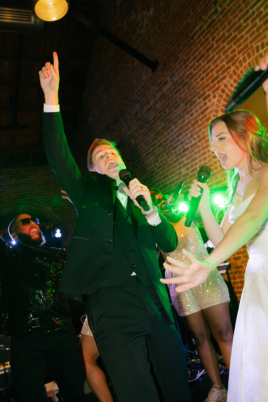 Bride and groom perform on stage, singing into microphones alongside the band during their reception celebration.