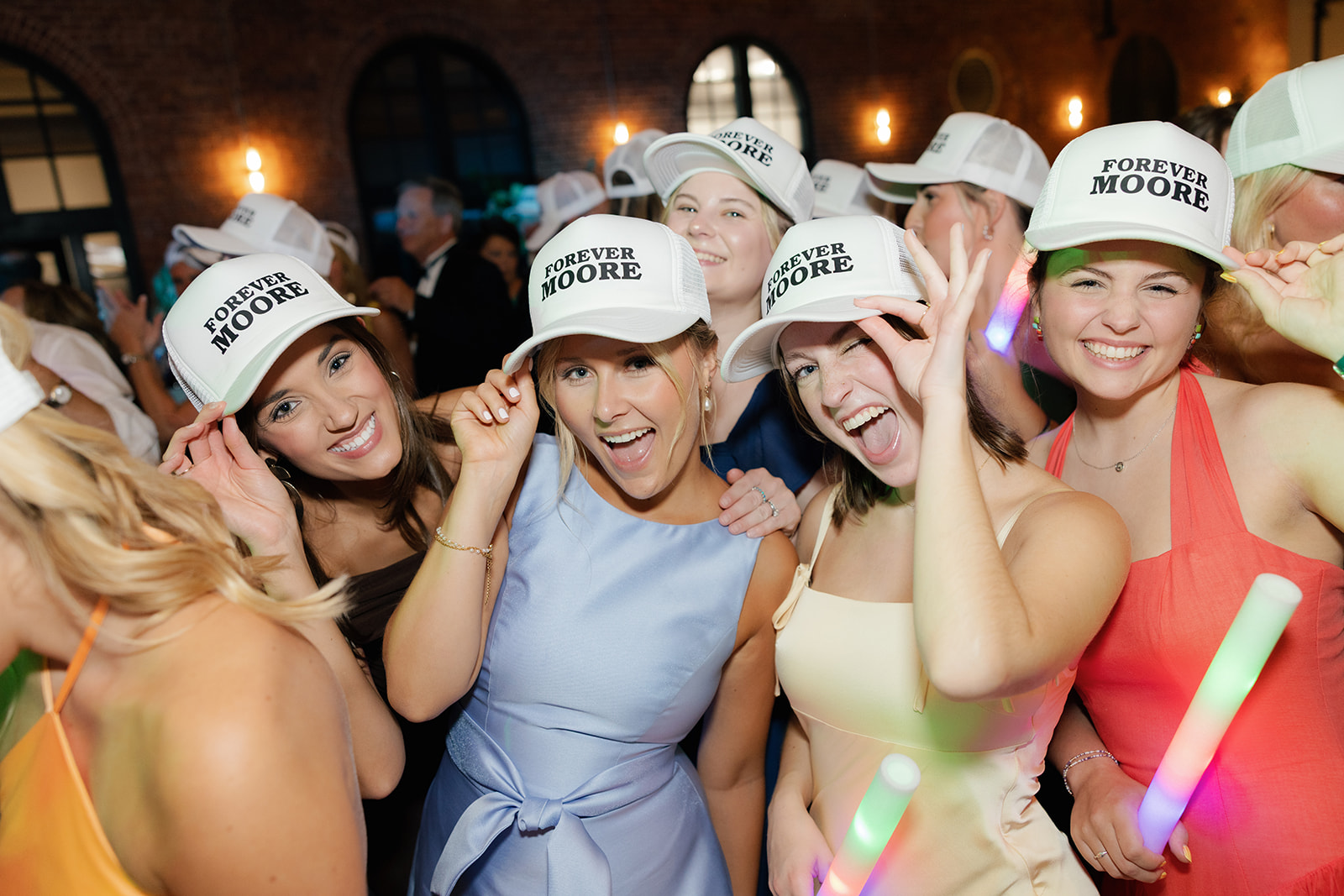 Smiling bridesmaids on the dance floor wearing matching “Forever Moore” hats and holding LED foam sticks.