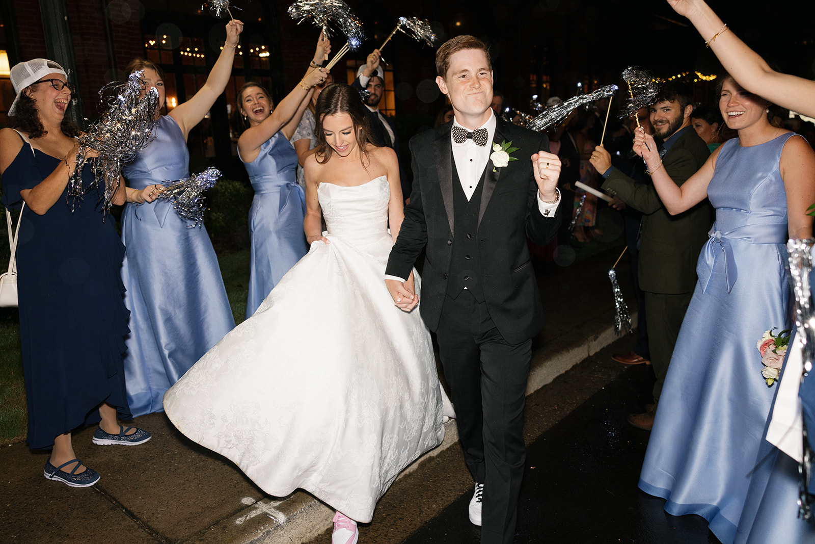 Bride and groom walk hand-in-hand through a cheering crowd at their wedding sendoff, bride wearing pink sneakers.