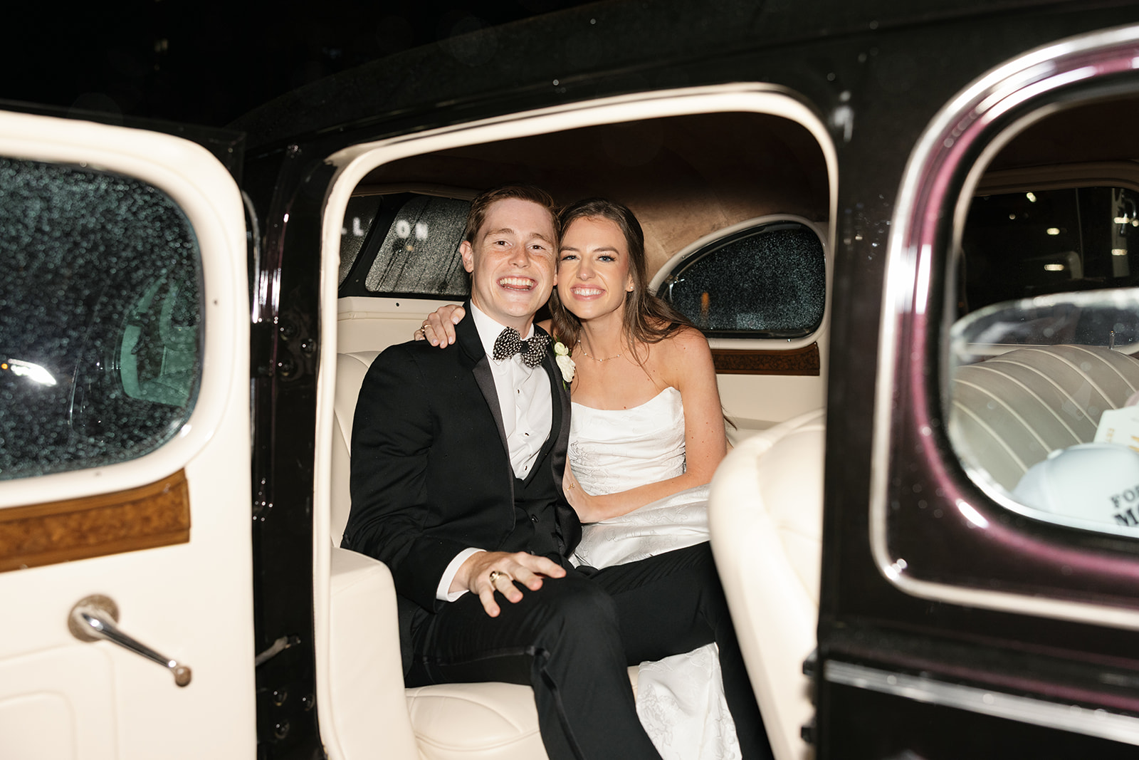 Bride and groom sitting in the back seat of a vintage getaway car, smiling and looking at the camera after their wedding.