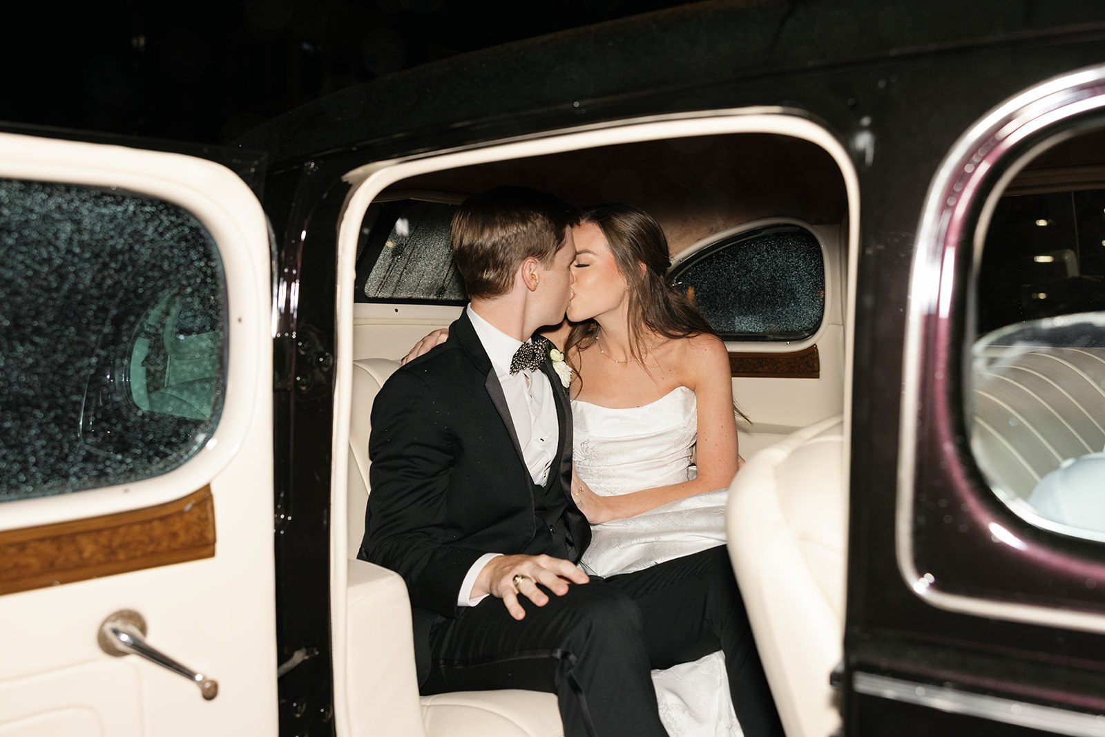Bride and groom share a romantic kiss in the back seat of their vintage getaway car to end their wedding celebration.