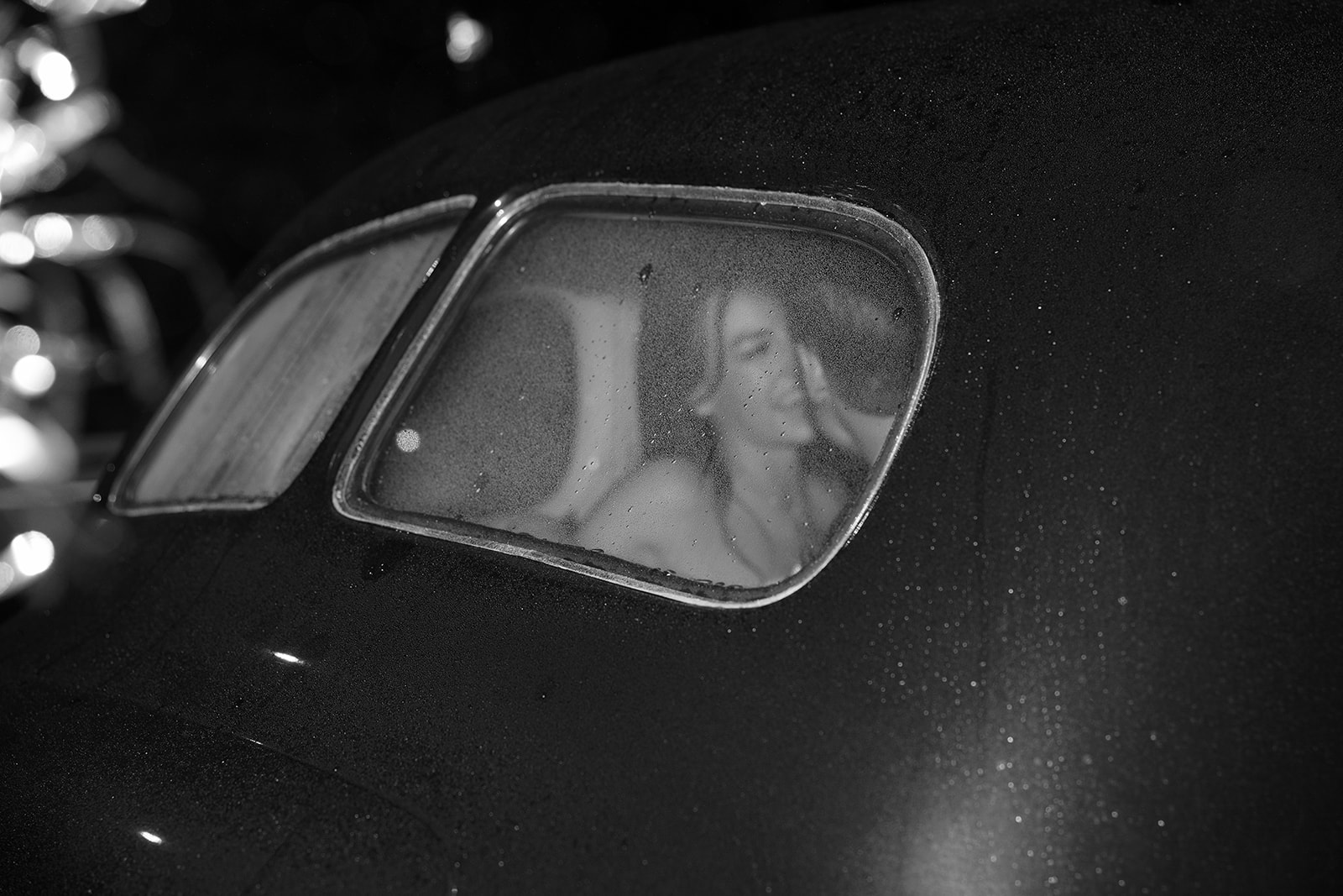 Moody black and white photo of bride and groom sharing a kiss through a rain-speckled car window on their wedding night.