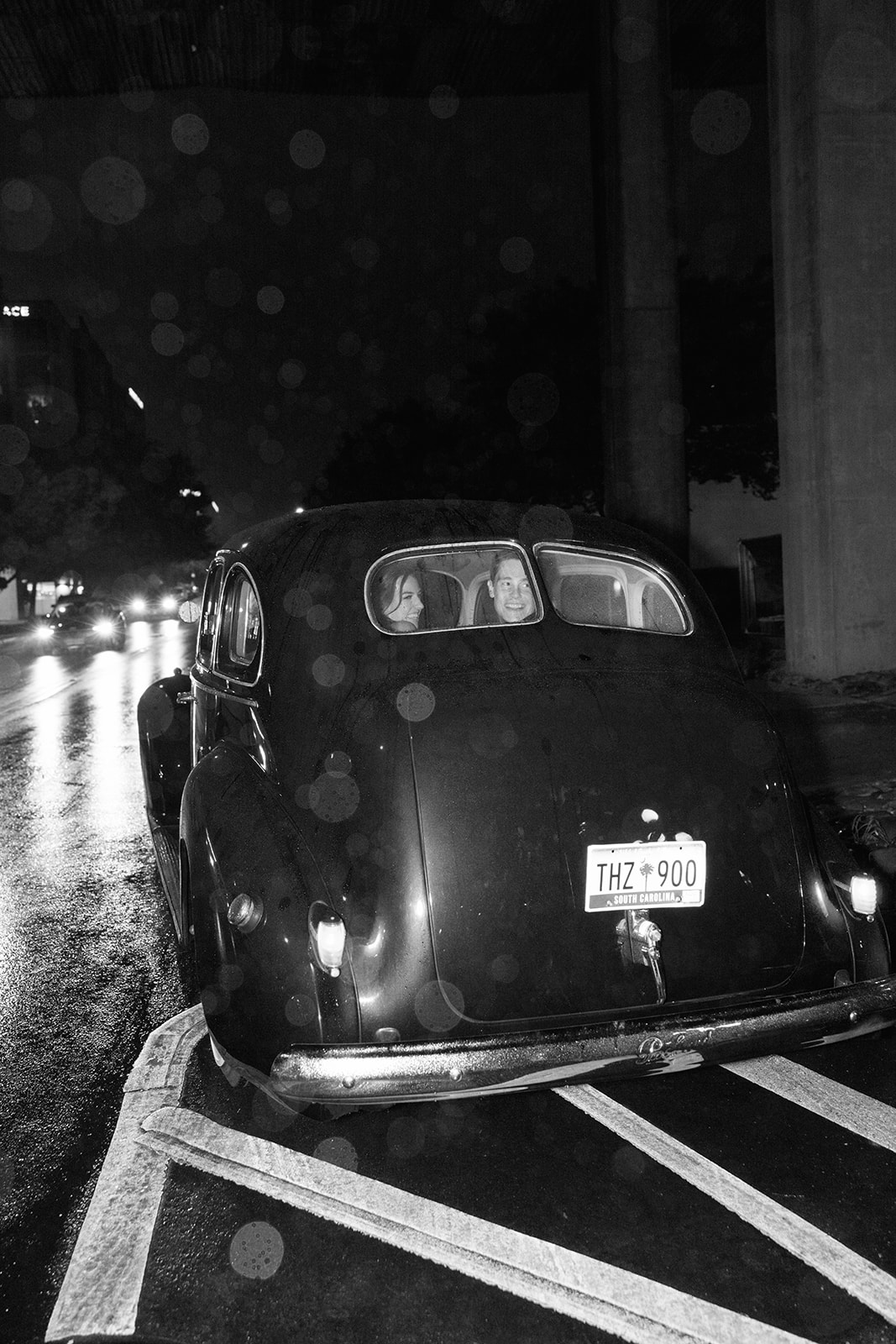 Black and white photo of newlyweds smiling through the rear window of a vintage getaway car on a rainy night.