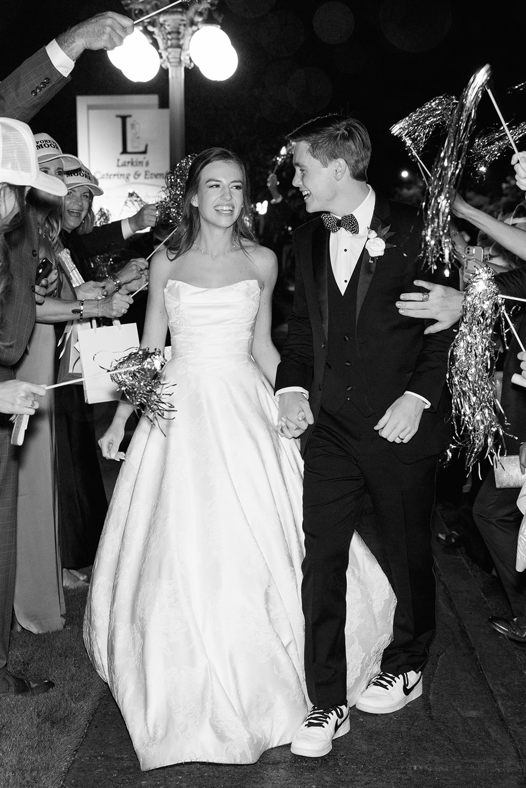 Black and white image of bride and groom laughing and holding hands during their wedding sendoff, lit by streetlights.